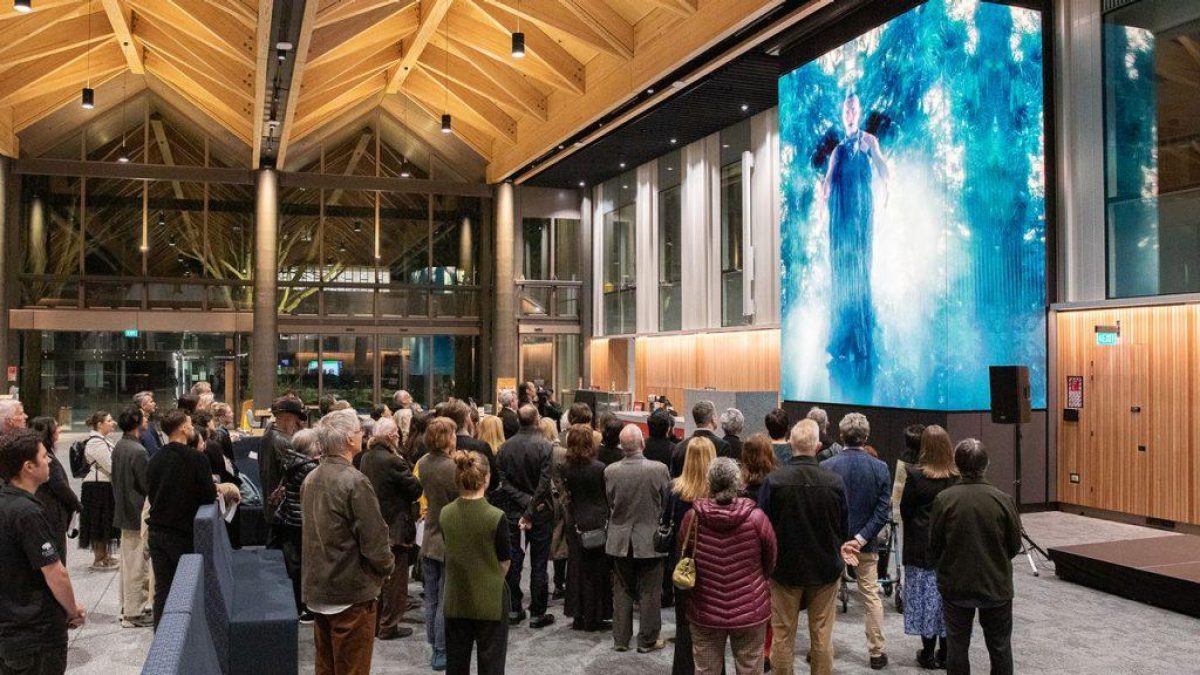 A large gathering of people viewing a towering, brightly lit video wall displaying abstract blue art in a modern public atrium with high wooden ceilings and glass walls.