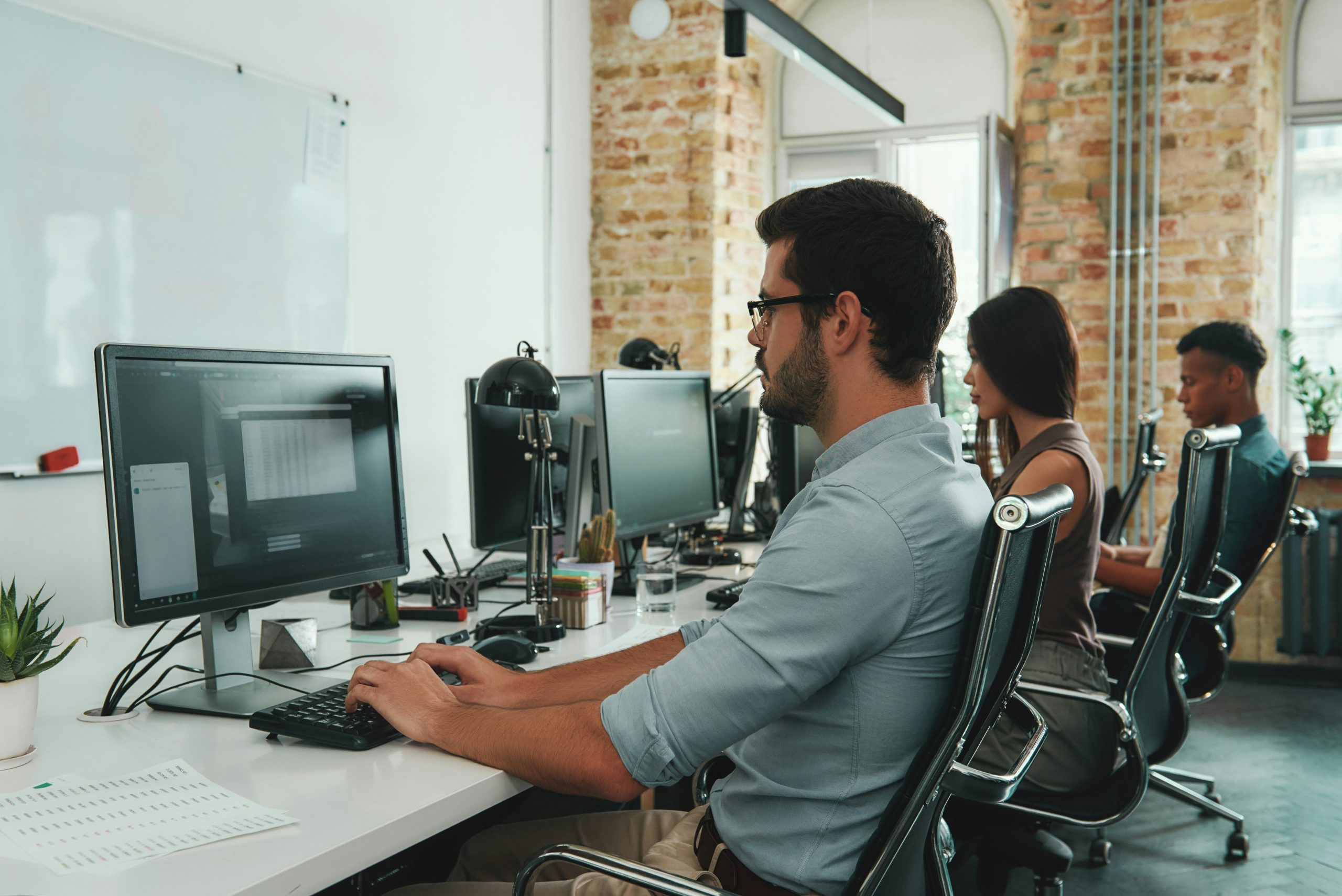 Three people sitting at desks in a modern office, working on desktop computers. Brick wall and whiteboard visible.
