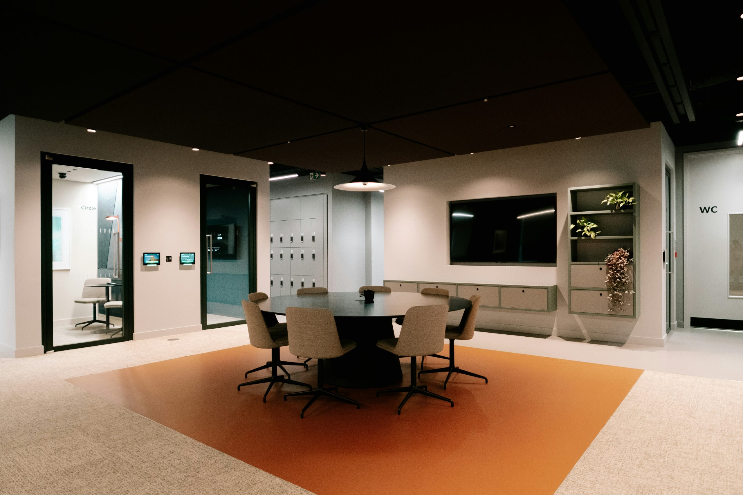 A modern office space with a round meeting table, chairs, wall-mounted screen, small meeting rooms, lockers and soft lighting, designed for quiet teamwork and collaboration.