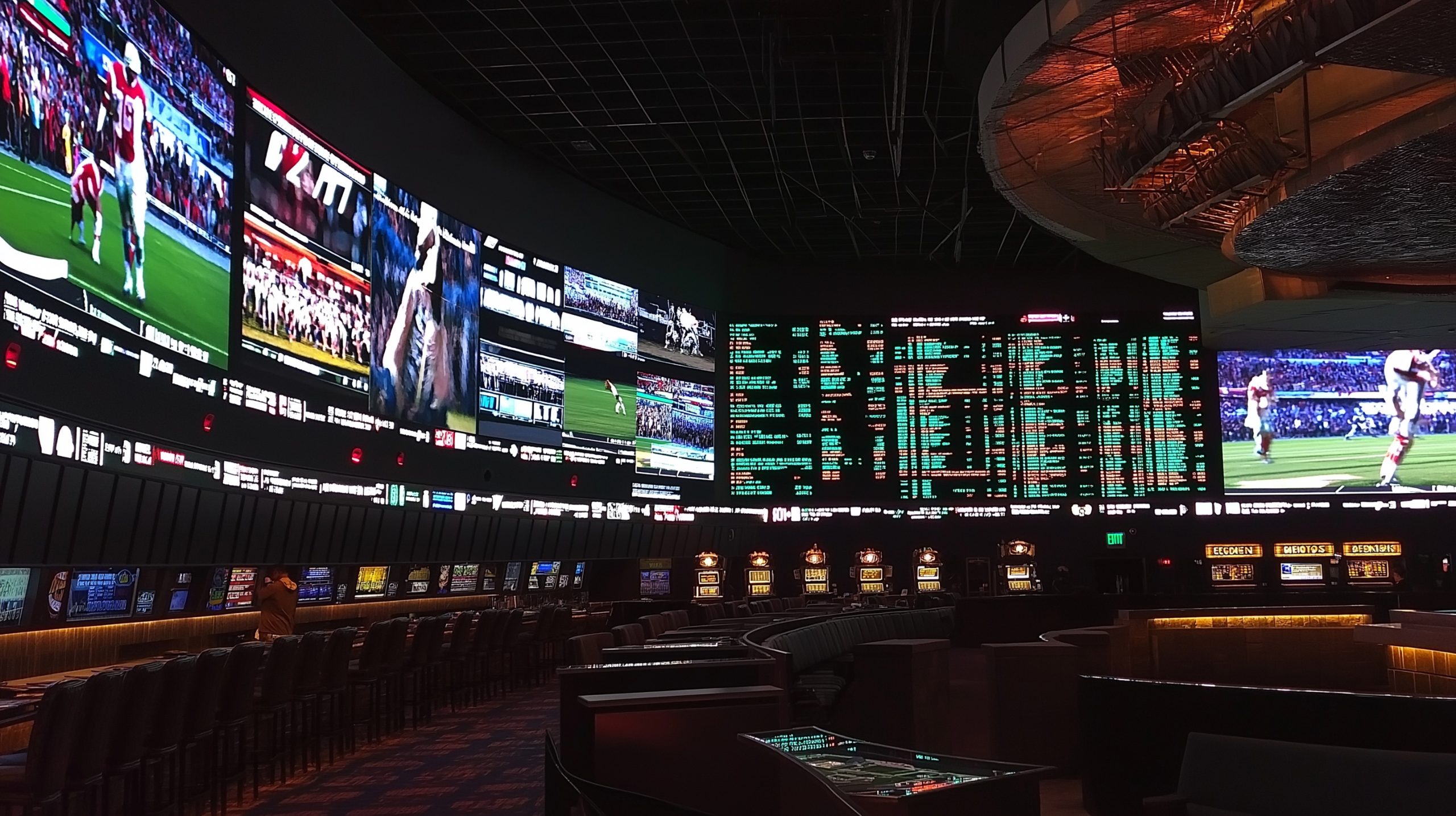 A dimly lit sports bar with numerous curved, interlocking screens displaying live sports and betting data. Seating for patrons is visible in the foreground.