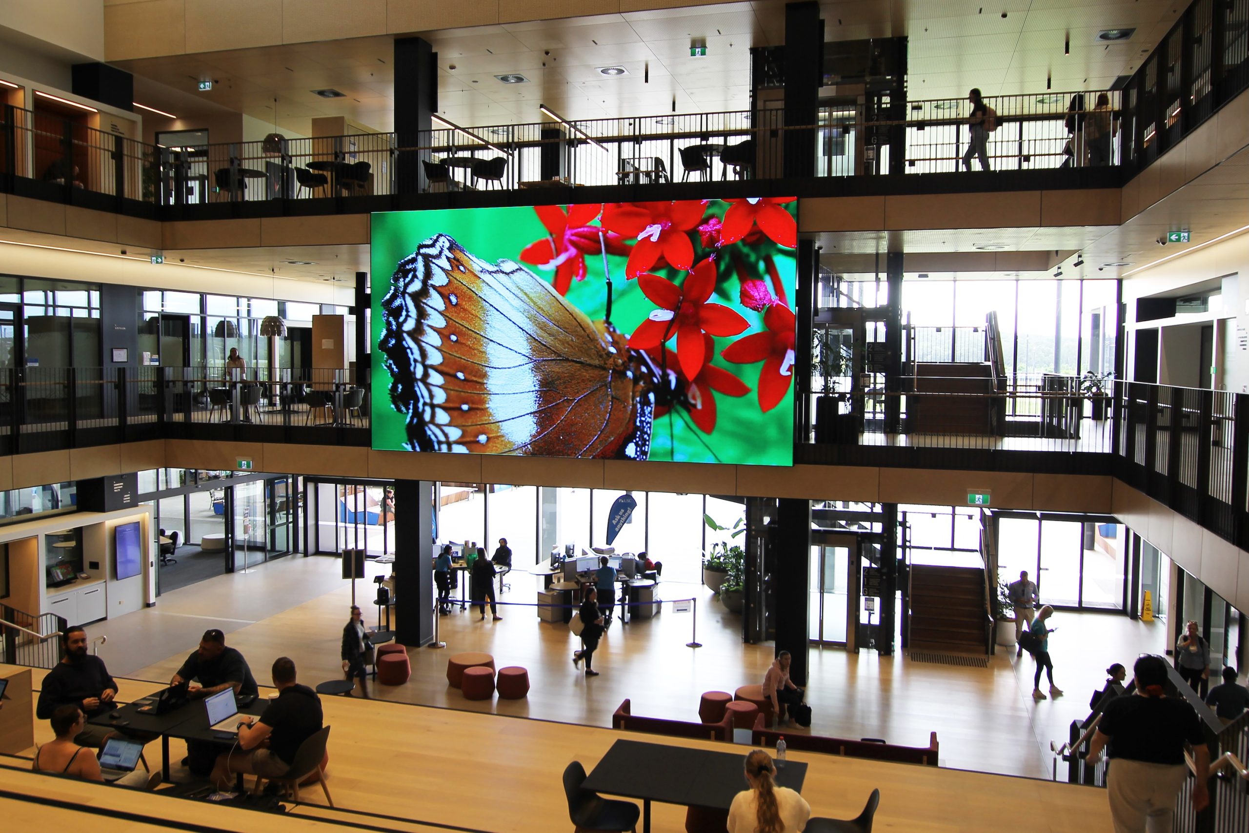 Large multi-panel LED screen displaying a butterfly and red flowers in a multi-story corporate building lobby with people circulating below.