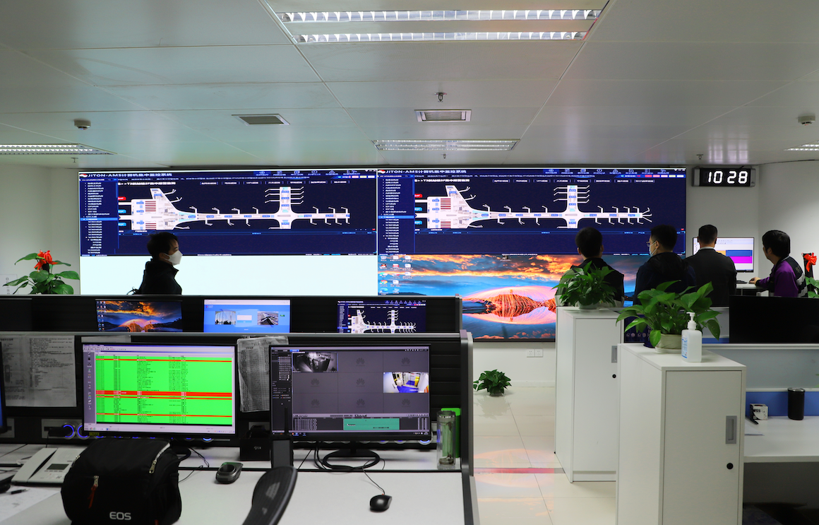 Control room with several large screens displaying data visualizations and monitoring charts. Two individuals are partially visible, observing the display wall.