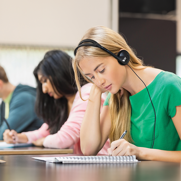 A student wears a Williams AV headset while writing in a spiral notebook during a class or language lecture.