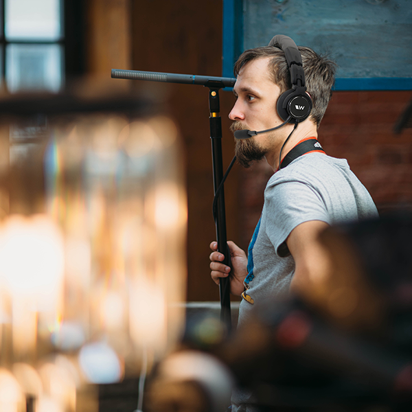 A production crew member wears a Williams AV headset with a boom mic and holds a microphone boom pole outdoors.