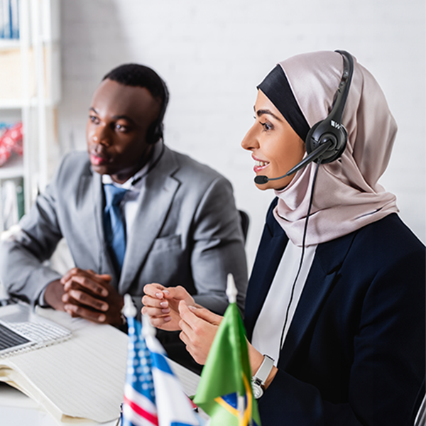 Two interpreters, one wearing a hijab, use headsets and microphones while seated at a desk with small flags.