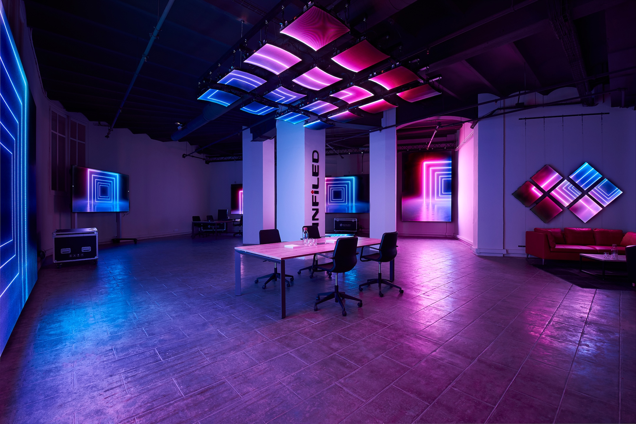 Open room with a central conference table, illuminated by pink and blue neon lighting from ceiling panels and vertical accents, creating an immersive, futuristic atmosphere.