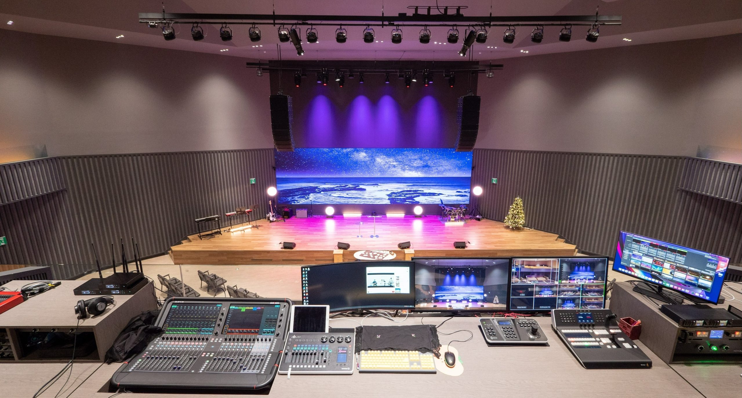 Interior shot of a modern auditorium with a stage, seating, professional lighting, and a large LED video wall displaying a starry landscape. Control systems are in the foreground.