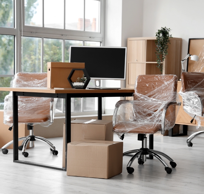 An office space partially packed for relocation, with chairs and computer monitors covered in plastic wrap. Cardboard boxes are stacked near a desk by a large window.