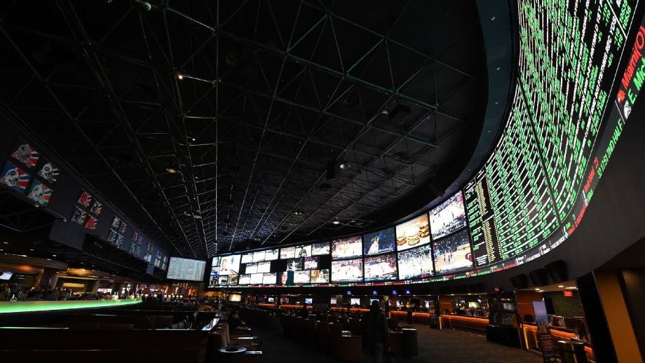 Interior of a dark sports book with tiered seating, featuring a massive curved LED screen displaying live betting odds, surrounded by multiple smaller screens showing games.