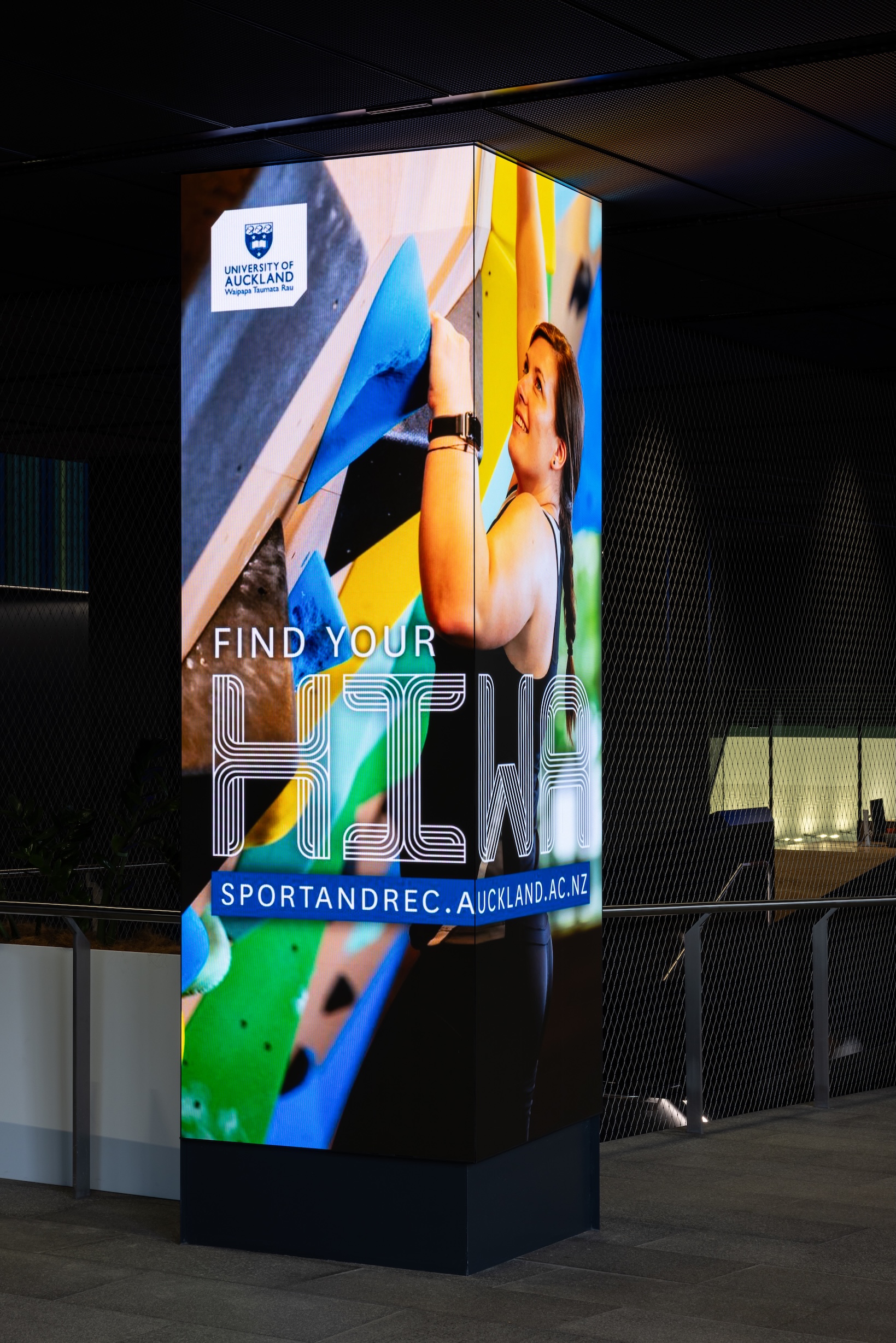 A tall, vertical digital display column featuring an advertisement for the University of Auckland Sport and Recreation center. The image shows a person rock climbing.