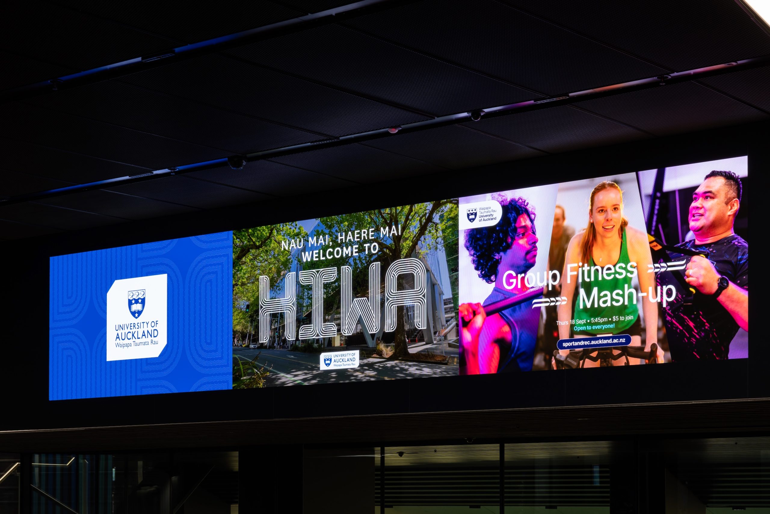 A large, illuminated indoor digital screen displays three distinct panels, including the University of Auckland logo, a welcome message in Māori and English, and an advertisement for a group fitness event.