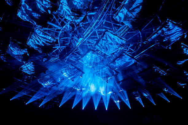 Blue spotlights shining brightly downward from a ceiling truss structure onto a dark area, with abstract shapes and fabric panels reflecting the intense light.