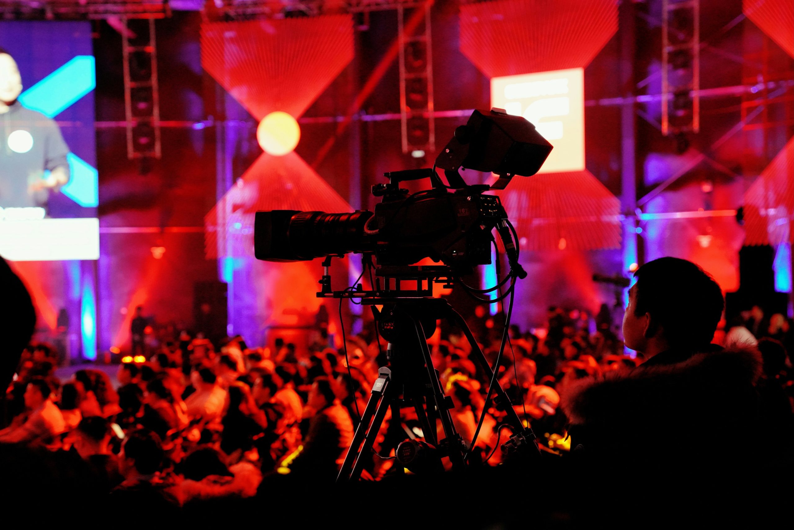 Digital video camera on a tripod recording a live event in a dark venue. The stage is lit with red and purple lights, and a large audience is visible in the foreground.