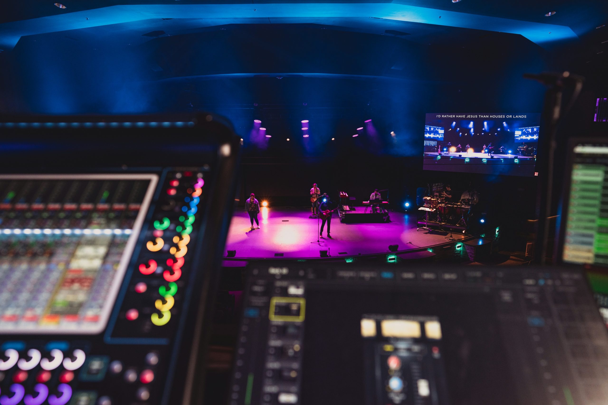 A professional sound mixing console in the foreground, with the colorful faders out of focus. In the background, a band performs on a stage lit with purple and blue lights.