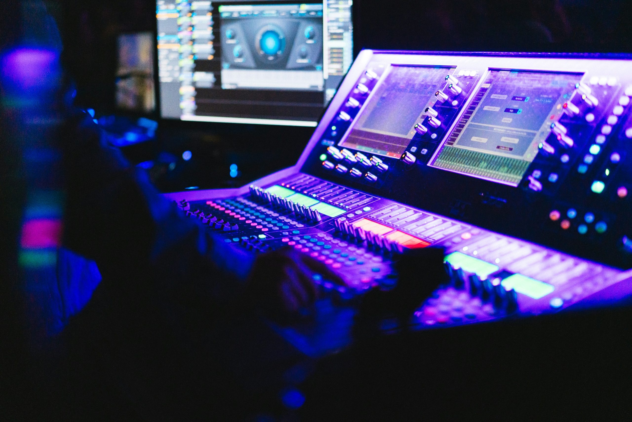 A professional sound mixing board in a dark environment, illuminated by purple and blue ambient light. Several digital screens on the console display audio equalization and level settings.