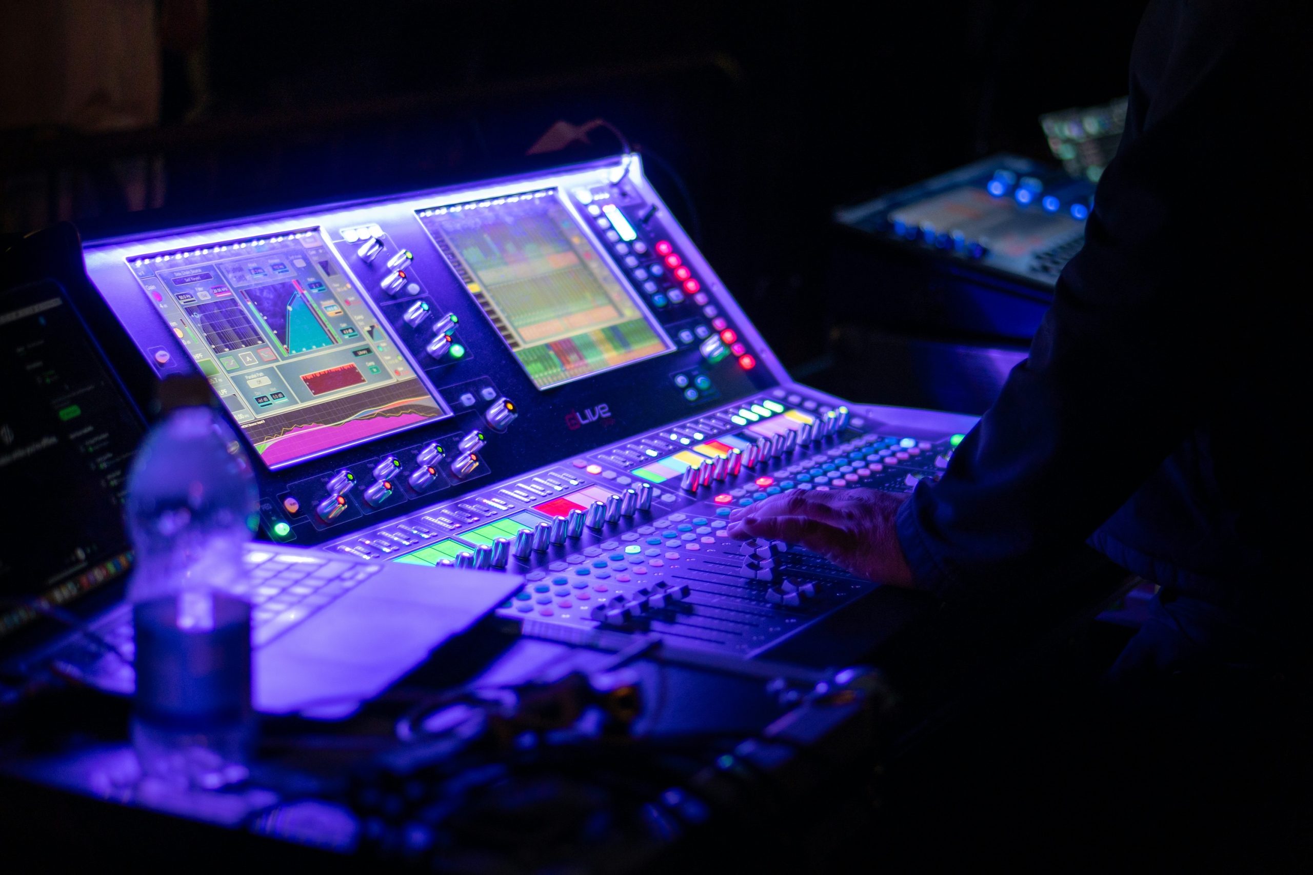 Audio technician operating a large digital sound mixing console illuminated with blue and purple light in a darkened control booth.